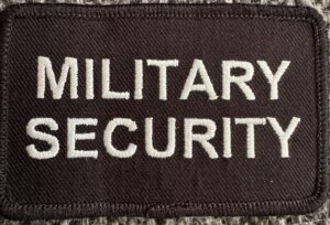 Militær sikkerhedsmærke med hvid broderet skrift.