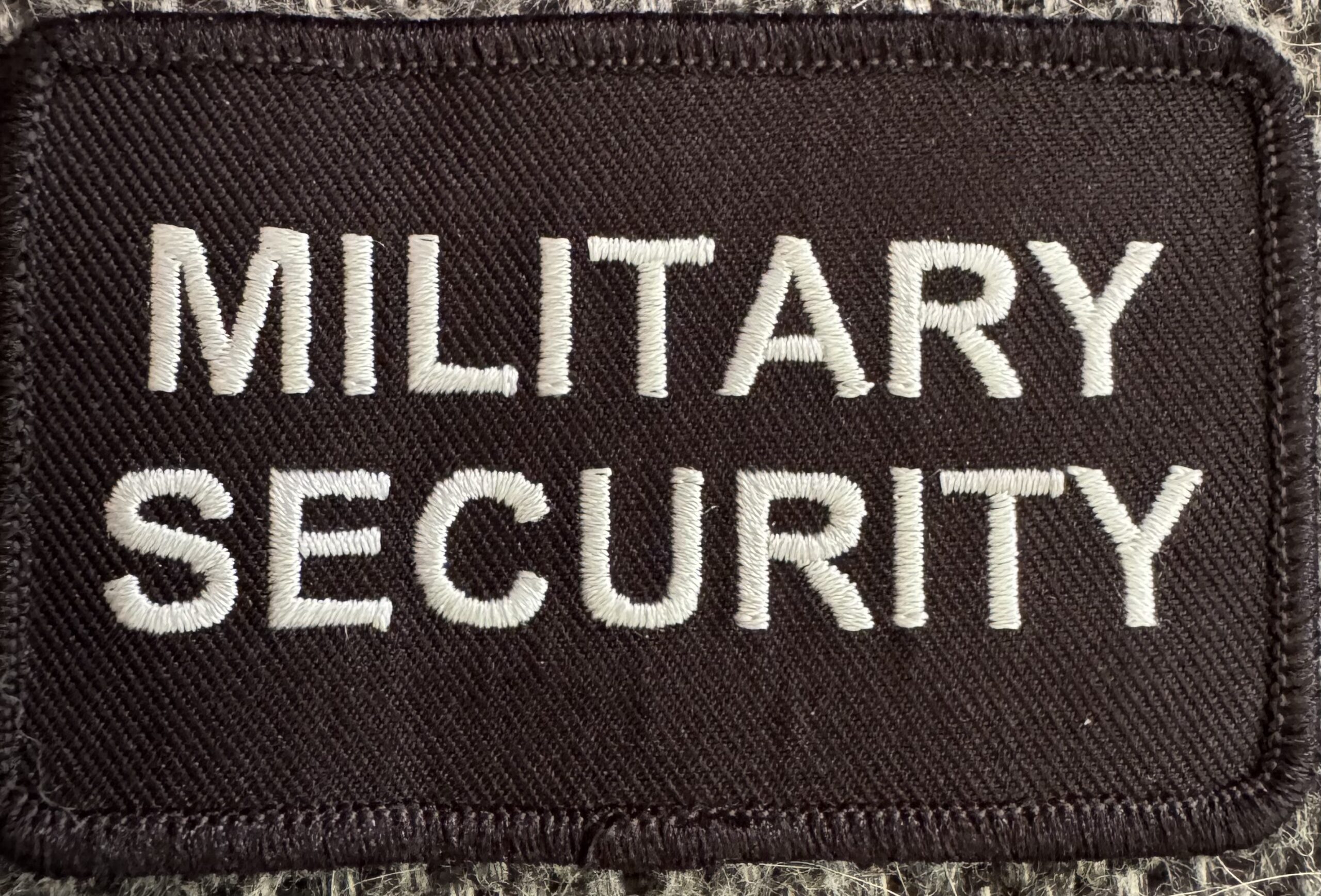 Militær sikkerhedsmærke med hvid broderet skrift.
